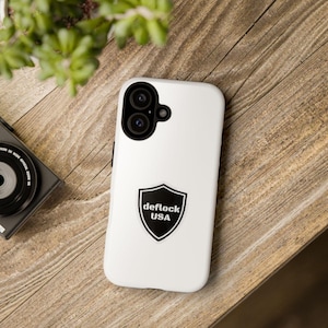 May include: A white smartphone case featuring a black shield-shaped emblem in the center. The emblem contains the white text "deflock USA" in a bold, sans-serif font. The case has a black camera module cutout at the top left corner.
