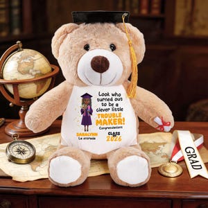 Puede incluir: Un oso de peluche marrón con birrete y diploma. La camiseta del oso presenta un gráfico de un graduado y el texto "Look who turned out to be a clever little TROUBLE MAKER!" y "Class of 2026".