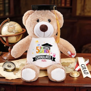 May include: A plush teddy bear wearing a graduation cap and a white bib. The bib reads "5th Grade Graduate 2026" with the name "Aizel" below. The bear is light brown with a dark brown nose and is sitting on a map.