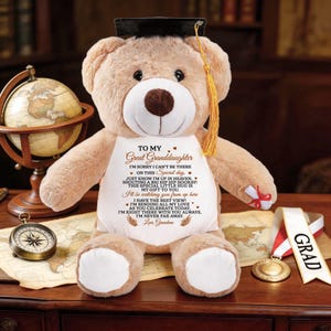 May include: A tan teddy bear wearing a graduation cap with a gold tassel. The bear has a white bib with a printed message to a great-granddaughter. A compass, globe, and medal are in the background.