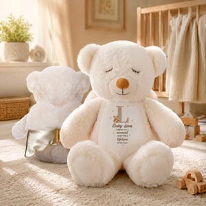 Könnte beinhalten: Ein cremefarbener Plüsch-Teddybär mit geschlossenen Augen und einer braunen Nase. Der Bär hat einen weißen Brustaufnäher mit dem Buchstaben L, einer kleinen Bärengrafik und dem Text Baby Liam, carried for a moment loved for a lifetime 03.06.2024.
