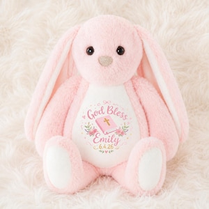 May include: A soft, light pink plush rabbit with long ears and dark eyes. The rabbit features a white belly patch embroidered with the text God Bless, the name Emily, and the date 6.4.26, surrounded by a pink book, flowers, and small hearts.