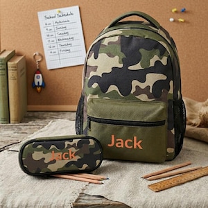 Op de afbeelding: Een rugzak en bijpassend etui met camouflageprint, beide voorzien van de naam Jack geborduurd in oranje. De rugzak heeft een voorvak met rits en zijvakken van gaas, terwijl het ovale etui een enkele ritssluiting heeft.