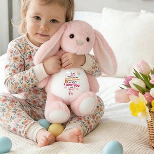 May include: A light pink plush bunny with long ears, held by a child. The bunny has a white belly with printed text and graphics. The text includes "I LOVE YOU" and "MIMI". The child is wearing floral pajamas and is surrounded by Easter eggs and flowers.