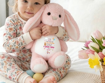 May include: A soft, light pink bunny toy with long ears, held by a child. The bunny features printed text and graphics on its white belly, including "I LOVE YOU" and "MIMI". The child wears floral pajamas and is surrounded by Easter eggs and a basket of flowers.