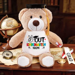 May include: A tan teddy bear wearing a graduation cap and a white bib. The bib reads "✌️ OUT KINDERGARTEN 2026 NAOMI" in colorful text. A gold tassel hangs from the cap. A compass, globe, and medal are in the background.