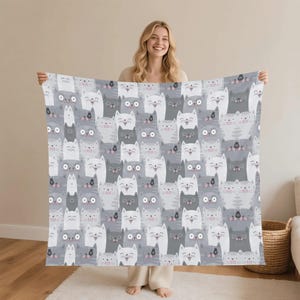 May include: A gray and white cat-themed blanket. The blanket features a repeating pattern of smiling cat faces in various shades of gray and white. The blanket is held up by a person.