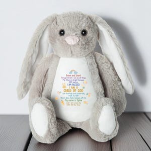 Puede incluir: Un conejito de peluche gris con orejas largas y una nariz rosada. El conejito tiene una barriga blanca con texto colorido que dice "I AM A CHILD OF GOD" y otras frases inspiradoras. El conejito está sentado sobre una superficie de madera.