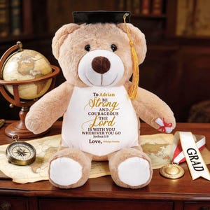 May include: A plush teddy bear wearing a graduation cap and a white bib with the text "To Adrian, Be Strong and Courageous, The Lord is with you wherever you go." A gold medal and a "Grad" ribbon are also visible.