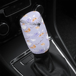 Peut inclure: Un manchon de levier de vitesse de voiture lavande avec un motif répété de théières, tasses à thé et pâtisseries blanches, orange et violettes. Les mots "tea time" sont écrits sur le manchon. Le manchon est cousu avec du fil noir.