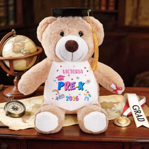 May include: A plush teddy bear wearing a graduation cap and a bib that says "Victoria PRE-K 2026". The bear is light brown with a white belly and foot pads. A graduation ribbon and medal are next to the bear.