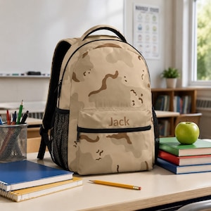 May include: A tan and brown camouflage print backpack featuring a front zippered pocket with the name Jack embroidered in brown thread. The backpack includes black mesh side pockets, a top handle, and adjustable shoulder straps for carrying.