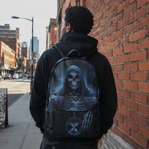 Peut inclure: Un sac à dos noir présentant une impression graphique détaillée d'un squelette à capuche tenant un sablier. Le motif est rendu dans des tons de bleu et de gris, couvrant le compartiment principal et la poche avant du sac avec des bretelles.