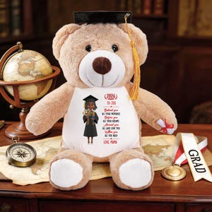 Puede incluir: Un oso de peluche beige con un gorro de graduación con una borla dorada. El oso tiene una camisa blanca con una ilustración impresa de un graduado y el texto "TO ZOE" y "LOVE, MAMA". Un globo terráqueo, una brújula y una medalla están en el fondo.