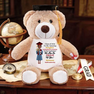 May include: A tan teddy bear wearing a graduation cap and gown. The bear has a white shirt with a graphic of a graduate and text that reads "Class of 2026 To My Amazing Daughter Congratulations On Your Graduation! We Are So Proud Of You! Maimuna Smith university."