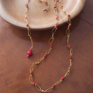 Puede incluir: Un collar largo que presenta una delicada cadena de color dorado con un intrincado diseño de alambre en bucles. La cadena está acentuada con pequeñas cuentas redondas de color rojo brillante espaciadas uniformemente, terminando en una cuenta roja más grande.