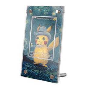Puede incluir: Una vitrina acrílica transparente que contiene una carta coleccionable de Pikachu. La carta presenta un Pikachu amarillo con un sombrero gris, sobre un fondo arremolinado azul y amarillo. La vitrina está sostenida por un soporte plateado.
