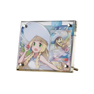 Könnte beinhalten: Ein transparentes Acryl-Display mit einer Pokémon-Sammelkarte, die Lillie zeigt, eine weibliche Figur mit blonden Haaren und einem weißen Hut. Die Karte ist von einem funkelnden blauen Hintergrund mit weißen Sternen umgeben. Der Text auf der Karte lautet "TRAINER'S - LILLIE".