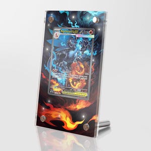 Puede incluir: Un marco de exhibición de acrílico transparente que contiene una tarjeta coleccionable Mega Charizard X EX. El marco presenta un fondo impreso con llamas azules arriba y naranjas abajo, asegurado por cuatro soportes metálicos plateados.