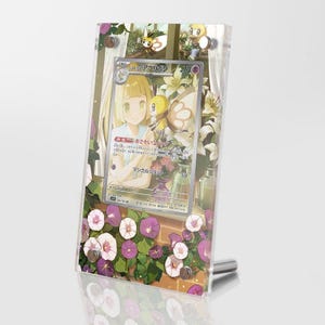 Puede incluir: Una vitrina de acrílico transparente con una carta de Pokémon que presenta a un personaje de cabello rubio con un Pokémon de tipo hada. La carta se titula "リーリエのプリズム". La vitrina está decorada con flores rosas y blancas.