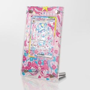 Puede incluir: Un expositor de plástico rosa y blanco con una carta de Pokémon que presenta un Pokémon hada azul claro y rosa. La carta tiene texto japonés que dice "Encanto de Hada" y "Sylveon".