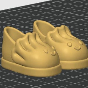 Op de afbeelding: Een paar lichtbruine instapschoenen met een gevormd, verhoogd ontwerp aan de voorkant dat lijkt op een knoedel met een klein, eenvoudig lachend gezichtje. De schoenen hebben een glad, effen oppervlak en een dikke, afgeronde zoolrand.