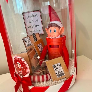 May include: A clear cylindrical display container with red and white striped trim containing a toy elf inside a red North Pole mailbox. The display includes miniature cardboard shipping boxes, a sign reading I couldn't resist... it was Elfazon Prime day!, and white foam beads.