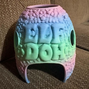 May include: A 3D printed, dome-shaped container with a textured, bumpy surface featuring a gradient of pastel pink, blue, and green colours. The front displays the embossed text "ELF DOH" in large, blocky letters, with circular openings on the sides.