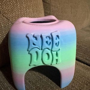 May include: A 3D printed tooth-shaped container featuring a pastel gradient of pink, blue, and green horizontal stripes. The front surface displays the embossed text "NEE DOH" in a bold, rounded font. The object has a hollow top opening.
