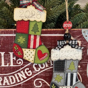 May include: Two hanging stocking ornaments. The left one features a red and green patchwork design with the year 2026 on the cuff. The right one has a black, gray, and white patchwork pattern with the name Lizzie and the year 2026 on a red tag.
