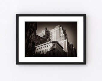 Könnte beinhalten: Schwarz-Weiß-Fotografie von Stadtgebäuden in einem Rahmen. Das Bild zeigt eine Perspektive von unten auf mehrere Gebäude, wobei die architektonischen Details hervorgehoben werden. Die Gebäude sind unterschiedlich hoch und gestaltet, was eine dynamische urbane Szene erzeugt.