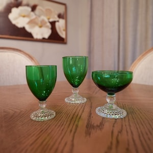May include: Three green glass goblets with clear stems and bases on a wooden table. The goblets are different sizes and shapes.
