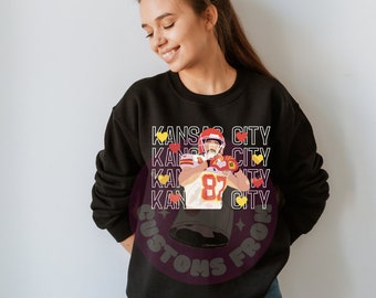 May include: Black sweatshirt with a graphic of a football player wearing a red and white jersey with the number 87. The text "KANSAS CITY" is repeated multiple times on the sweatshirt with yellow hearts.