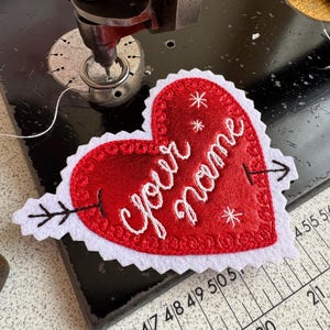Puede incluir: Un parche de tela rojo en forma de corazón con bordes festoneados blancos, que presenta el texto bordado "your name" en cursiva blanca, acentuado por un diseño de flecha negra y motivos de estrellas blancas, sobre una base de máquina de coser negra.