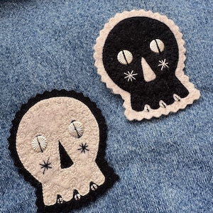 May include: Two skull-shaped patches. One is black with a light beige scalloped border, silver button eyes, and embroidered details. The other is light beige with a black scalloped border, silver button eyes, and embroidered details. Both are on a blue denim background.