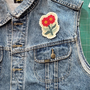 May include: A vintage denim vest with a floral patch. The vest is a light blue wash with a classic collar, button-up front, and two front pockets. The floral patch features red flowers, green leaves, and a cream-colored border. The vest has the "Lee" brand label.