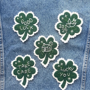 Puede incluir: Una chaqueta vaquera adornada con cinco parches de trébol verde. Cada parche presenta texto y detalles en blanco, incluyendo frases como "Good Luck" y "Lucky You". Los parches están bordados con hilo blanco y tienen un borde blanco.