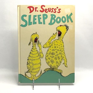 May include: A hardcover copy of "Dr. Seuss's Sleep Book" with a light beige cover. The title is in red and teal lettering. The cover illustration features two yellow, furry creatures yawning widely, set against a light beige background and a green base.