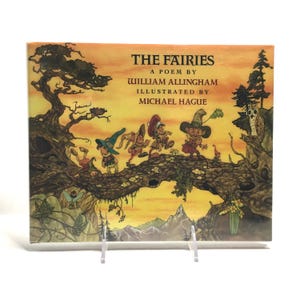 May include: A hardcover book titled "The Fairies" by William Allingham, illustrated by Michael Hague. The cover features a whimsical illustration of fairies on a tree branch against a sunset backdrop. The book is displayed on a clear stand.