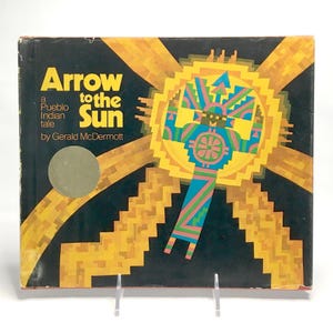 May include: Vintage hardcover book titled "Arrow to the Sun" by Gerald McDermott. The cover features a geometric design of a sun with a central figure in shades of yellow, blue, green, and pink against a black background. A Pueblo Indian tale.
