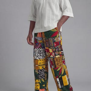 May include: A white short-sleeve shirt with a textured vertical stripe pattern, paired with patchwork print pants. The pants feature a vibrant mix of colors and patterns, including yellow, red, green, and black. Black sandals complete the outfit.