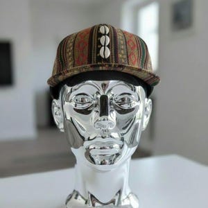 May include: A patterned baseball cap with a brown, red, and green design, adorned with three white shell-shaped embellishments. The cap sits atop a silver, metallic bust of a human head.