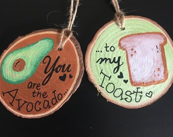 May include: Two wooden ornaments with painted designs. One ornament is brown with a green avocado and the text "You are the Avocado..." The other ornament is green with a white slice of toast and the text "...to my Toast".