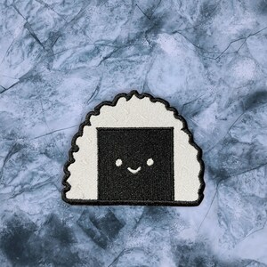 May include: A white and black embroidered patch of a smiling onigiri, a Japanese rice ball.