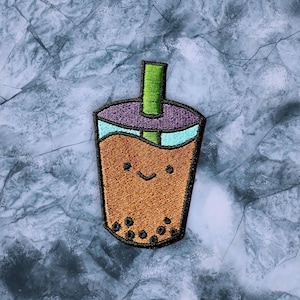 Peut inclure: Un patch brodé représentant un gobelet de bubble tea de dessin animé avec une paille verte, des accents violets et bleus et un visage souriant. Le gobelet est rempli de liquide brun et a des points blancs représentant des boba.