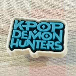 Pu&ograve; includere: Un charm per scarpe bianco e blu con la scritta "KPOP DEMON HUNTERS" in un carattere audace e stilizzato. Il testo &egrave; di colore blu brillante, delineato in nero e su sfondo bianco.