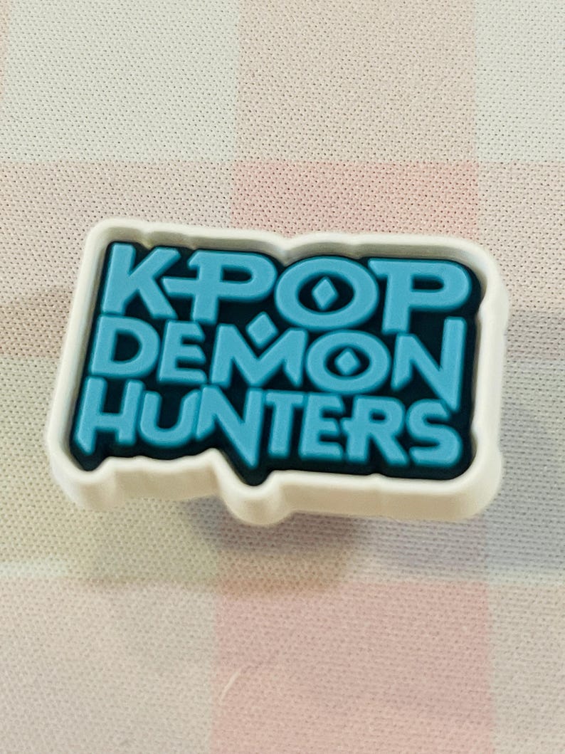 Pu&ograve; includere: Un charm per scarpe bianco e blu con la scritta "KPOP DEMON HUNTERS" in un carattere audace e stilizzato. Il testo &egrave; di colore blu brillante, delineato in nero e su sfondo bianco.
