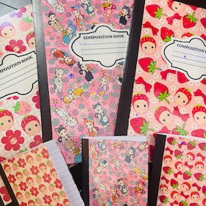 May include: Composition notebooks with vibrant covers. One notebook displays a pink background with strawberries and bows. Another features cartoon characters and flowers on a pink background. The notebooks are labelled "COMPOSITION BOOK" on the front.