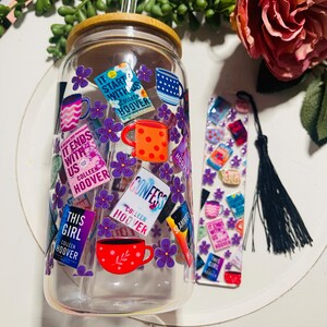 Op de afbeelding: Een doorzichtige glazen beker met een bamboe deksel en een rietje. De beker is versierd met kleurrijke stickers met boekomslagen en titels van Colleen Hoover, waaronder "It Ends With Us", "Confess", "This Girl" en "Slammed". De beker wordt geleverd met een bladwijzer met een vergelijkbaar ontwerp.