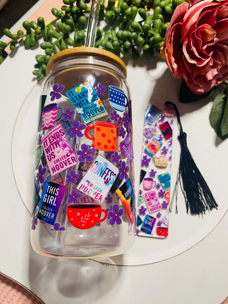 Op de afbeelding: Een doorzichtige glazen beker met een bamboe deksel en een rietje. De beker is versierd met kleurrijke stickers met boekomslagen en titels van Colleen Hoover, waaronder "It Ends With Us", "Confess", "This Girl" en "Slammed". De beker wordt geleverd met een bladwijzer met een vergelijkbaar ontwerp.
